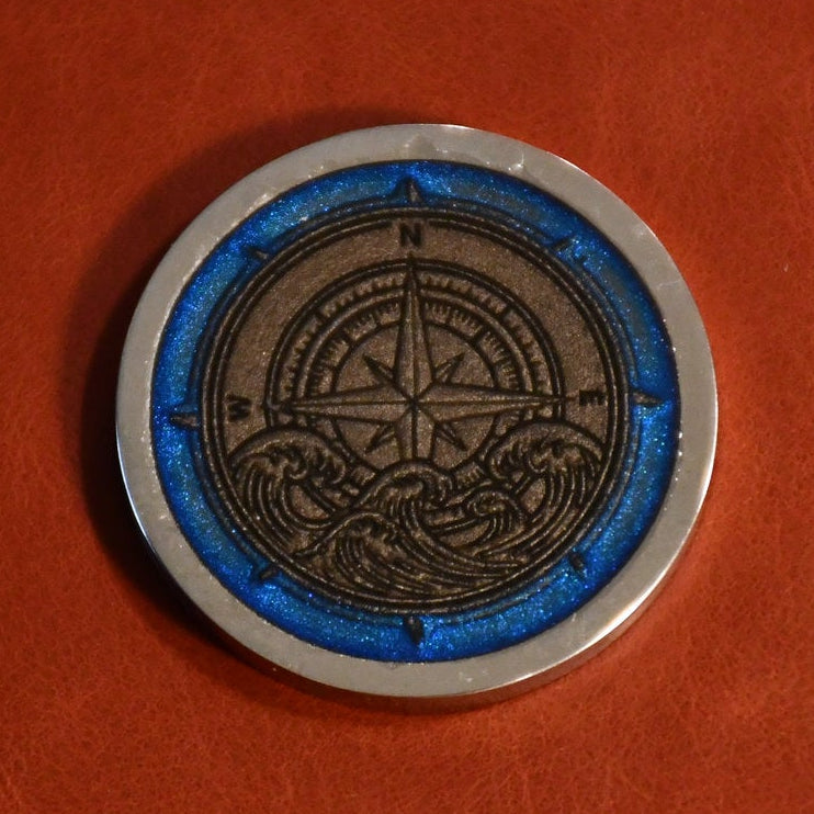 A circular silver-toned coin or medallion with an engraved compass rose design and blue accents, placed on a textured reddish-brown surface.